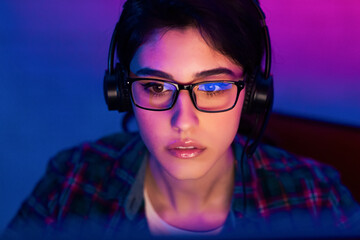 A young woman with dark hair, wearing eyeglasses and headphones, is focused on a computer screen in front of her. The image is lit by blue and purple lights © Prostock-studio