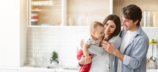 A happy family stands in a modern kitchen, with the mother holding their baby while the father smiles and looks at the child.