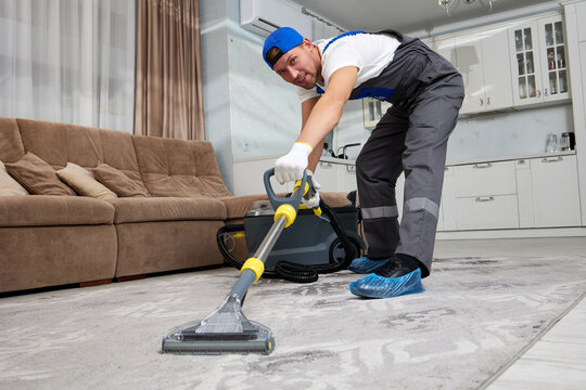 A smiling cleaning company employee in overalls cleans the customer's carpet with a professional cleaning vacuum cleaner.