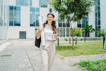 One young caucasian woman student going to university while listening to music on wireless headphones and holding papers and coffee	