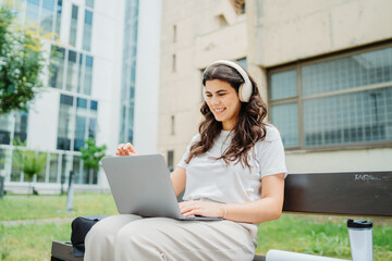 One beautiful young caucasian woman student sitting on bench near university listening to music and studying on laptop