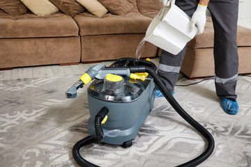 Professional cleaner in protective gear pouring water into cleaning vacuum. Person is wearing gloves, mask, and uniform, ensuring safety and attention to detail.
