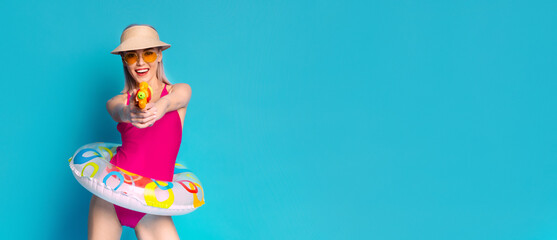 A young woman is wearing a pink swimsuit and a straw hat, standing with a bright yellow water gun in her hand. She is holding an inflatable ring in front of her and is smiling broadly.