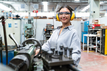 A young woman wearing safety glasses and ear protection is operating a lathe in a metalworking shop. looking at the camera and smiling.