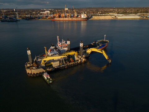 Harbour maintenance with backhoe dredger in Southampton Docks, UK. Hydraulically driven excavator on pontoon dredging. Aerial view at golden hour. Cargo ship at distance.
