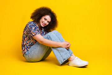 Full body photo of attractive young woman hugging knees girlish pose dressed stylish colorful clothes isolated on yellow color background