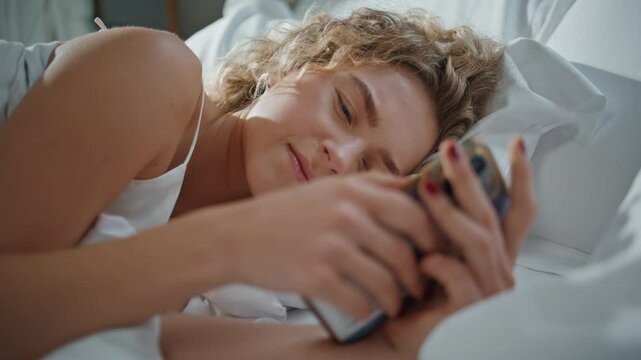 Woman typing mobile phone in bed closeup. Sleepy curly girl checking cellphone