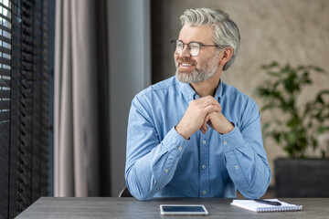Thoughtful mature office worker sitting at his desk, looking out the window and contemplating. He...