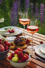 Romantic table decor for a loving couple on the blooming meadow with purple lupines. Two glasses of wine, flowers in a vase, silverware, fruits, wooden furniture and picnic basket. Sunset, golden hour