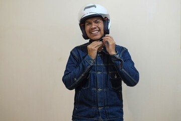 A Southeast Asian man wearing denim jacket and fastening his motorbike helmet with cream background, Positive person.