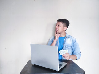 Mature asian man is thinking something while holding money in front of his laptop