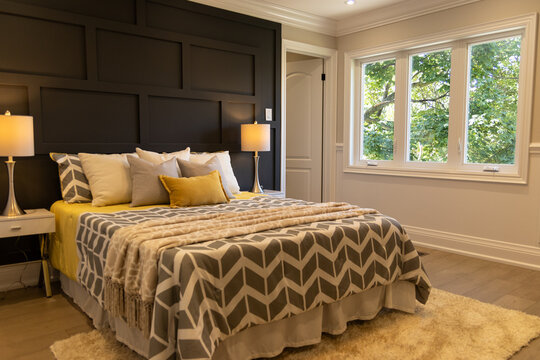 Brightly-lit bedroom - large bed with geometric-patterned comforter and yellow accents - nightstands with lamps - window view of greenery. Taken in Toronto, Canada.