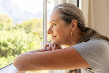 Smiling mature woman gazing out window, enjoying peaceful moment at home