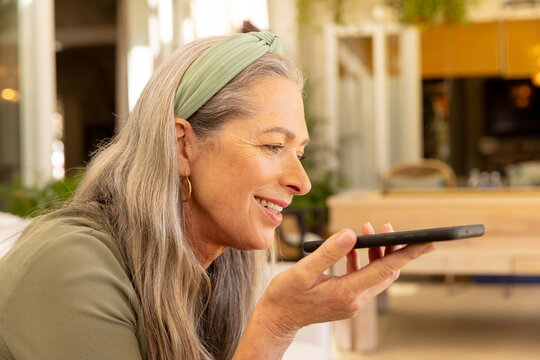 Smiling mature woman using smartphone for voice message at home, enjoying technology
