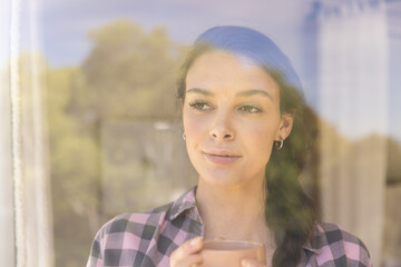 Young woman holding coffee cup, looking outside window, enjoying peaceful moment