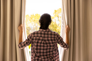 Opening curtains, woman in plaid shirt looking outside at sunny day