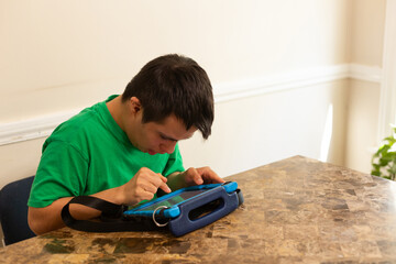 A young man with down syndrome uses a tablet at the dining room table. He wears a green shirt.