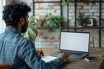 Fototapeta premium Over the shoulder view of Indian man working on a laptop with blank screen for mockup at modern office desk taking notes during virtual meeting or online training