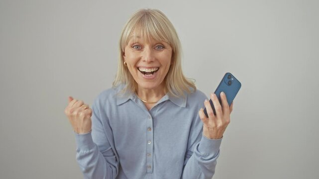 Cheerful young caucasian woman, blonde, beaming with excitement as she gleefully celebrates her triumphant victory. holding her smartphone, with arms raised, against an isolated white background.