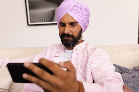 Using smartphone, man in pink turban sitting on couch at home - Powered by Adobe
