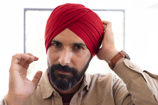 Adjusting red turban, man with beard looking confidently at camera indoors