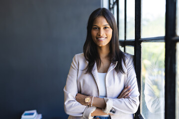 Smiling businesswoman standing with arms crossed by office window, looking confident