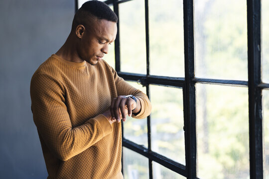 Checking smartwatch, man standing by window in casual sweater, focusing on time