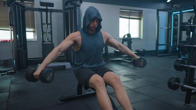Athletic Man Doing Side Dumbbell Raises Sitted on Bench in Empty Gym, Wearing Shorts and Tank Top
