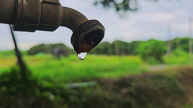 
A close-up view of the final drop of water falling from a rusty tap, highlighting the pressing issue of water scarcity against a blurred, green landscape backdrop.