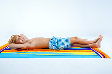 Happy smiling preteen boy in blue plaid swim trunks lying on his back comfortably on a colorful beach towel