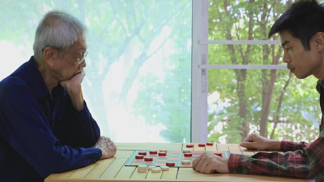 Senior asian father and son are enjoy playing checkers board game at home for leisure activities hobby and preventing from alzheimer symptom illness