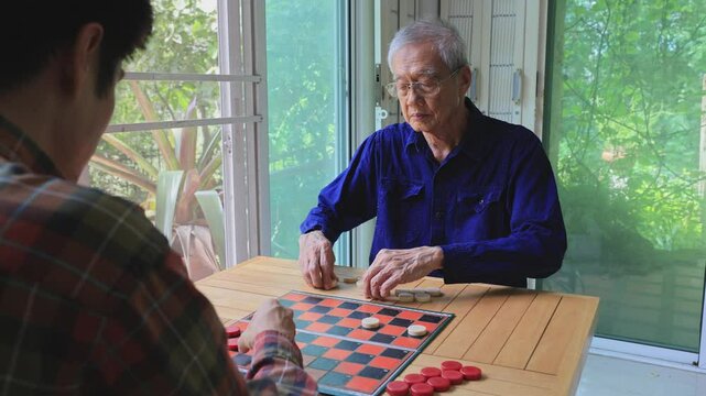 Senior asian father and son are enjoy playing checkers board game at home for leisure activities hobby and preventing from alzheimer symptom illness