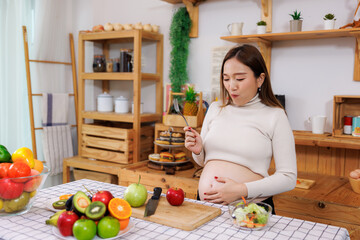 Pregnant asian woman standing eating salad in kitchen and hand touching her belly,