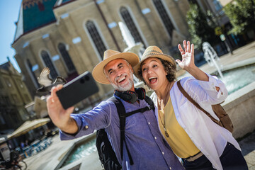 Elderly married couple exploring new city. Taking selfie in the centre. They are wearing hats and backpacks.
