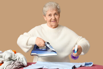 Happy senior woman with laundry and spray bottle ironing clothes on brown background
