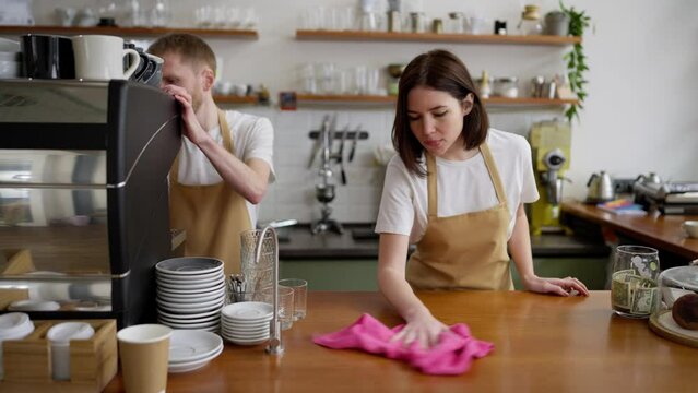 A brunette girl in a yellow apron cleans the counter with a pink rag while preparing for a working day with a barista guy in a cafe