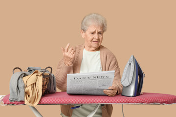 Displeased senior woman with basket of laundry and iron reading newspaper on brown background