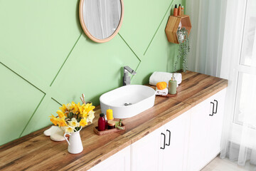 Modern sink, bath supplies and flowers on wooden chest of drawers near green wall in bathroom