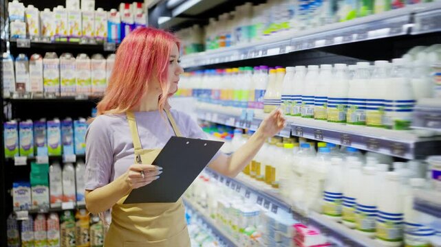 A girl supermarket worker with pink hair holds a tablet in her hands and takes inventory of dairy products on a shelf in a supermarket