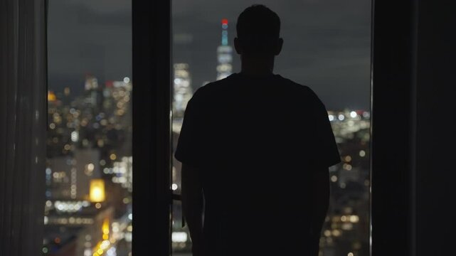 Lonely man standing with back to camera in silhouette against metropolitan cityscape at night watching the city in sad emotional look. His hand touching the glass window of hotel apartment or office