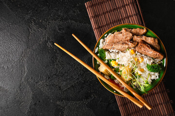 Bowl of tasty rice, chicken, vegetables, bamboo mat and chopsticks on dark background