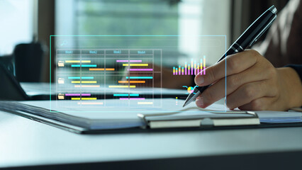 Hand holding pen with a holographic project management chart overlay in an office. Business analytics and planning concept.