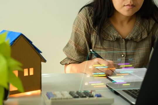 Woman using a futuristic holographic interface to analyze data while working from home. A small model house and calculator are on the desk.