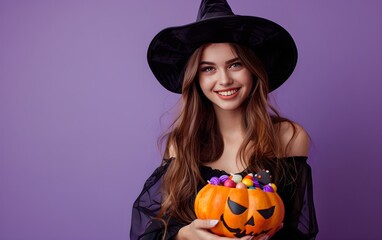 Young woman in witch costume with pumpkin basket of candies on purple background. Studio portrait of beautiful smiling girl. Halloween celebration, trick or treat concept. Banner with copy space