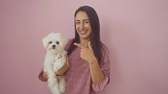 Young smiling hispanic woman pointing with finger hand while standing with her happy dog over isolated pink background