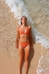 Fototapeta premium A woman in an orange bikini lies on the beach with her eyes closed, sunbathing and letting the gentle waves touch the sand around her, creating a peaceful scene.