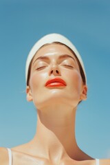 A woman in a white outfit and a sun hat basks in the sunny beach weather. Her red lipstick shines against her glowing skin while she enjoys the warmth and light with closed eyes.