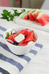 Summer salad with watermelon, feta cheese, olives and mint in a white bowl on a white wooden table. Concept of healthy eating. Vegan, vegetarian. Vertical orientation. Selective focus.