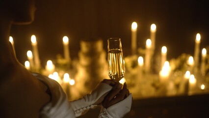 Close-up of a young woman's hand holding a glass of champagne at Christmas, illuminated by Christmas lights. In hands is a glass of champagne against a background of burning candles.