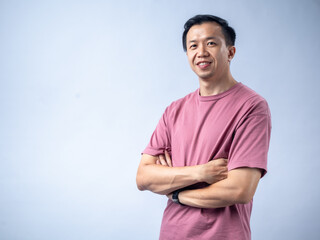 An Asian man in a pink shirt stands confidently with his arms crossed, smiling warmly at the...